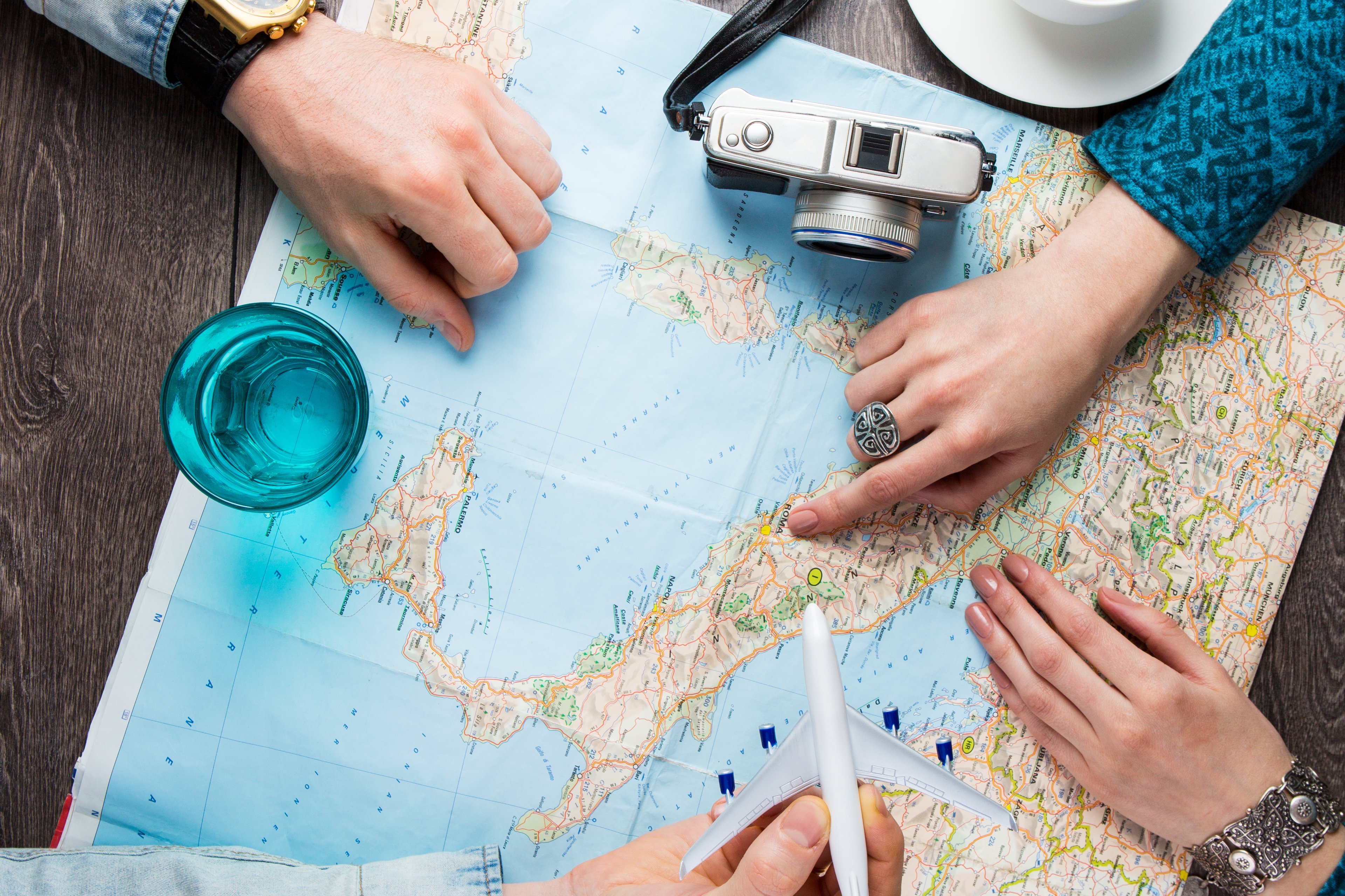 Landkarte mit zwei Personen die auf die Karte schauen und eine Reise planen, daneben stehen ein Kaffee, ein Fotoapparat und ein Glas Wasser