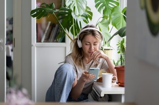 Frau, die Podcast hört mit drahtlosen weißen Kopfhörern, mit Handy in der Hand und einer Tasse Tee auf dem Tisch