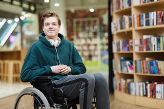 Junger Mann sitzt in einem Rollstuhl in einer Bibliothek und lächelt in die Kamera
