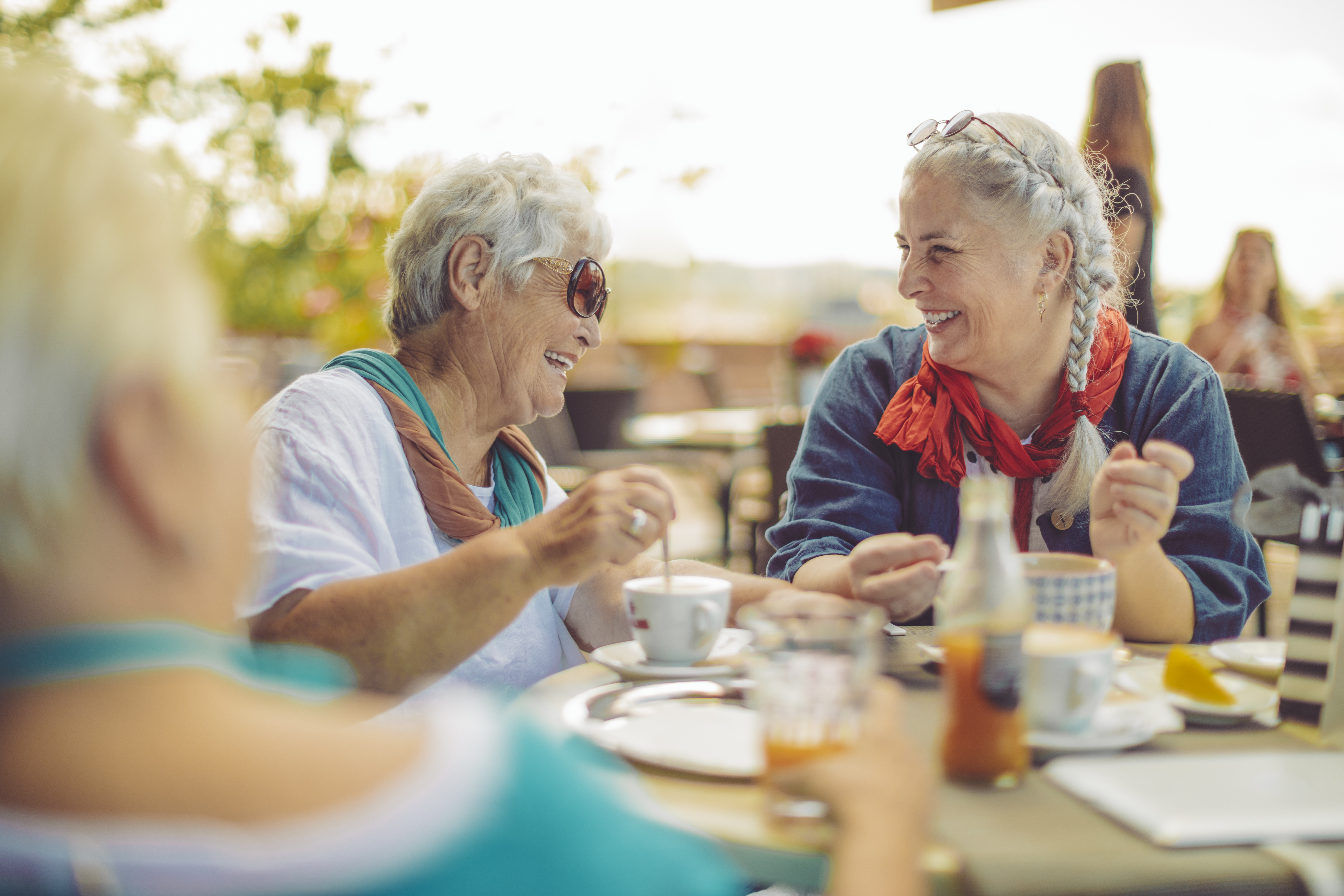 zwei Frau mittlere Alters sitzen lachend am Tisch im Freien und unterhalten sich bei einer Tasse Kaffee